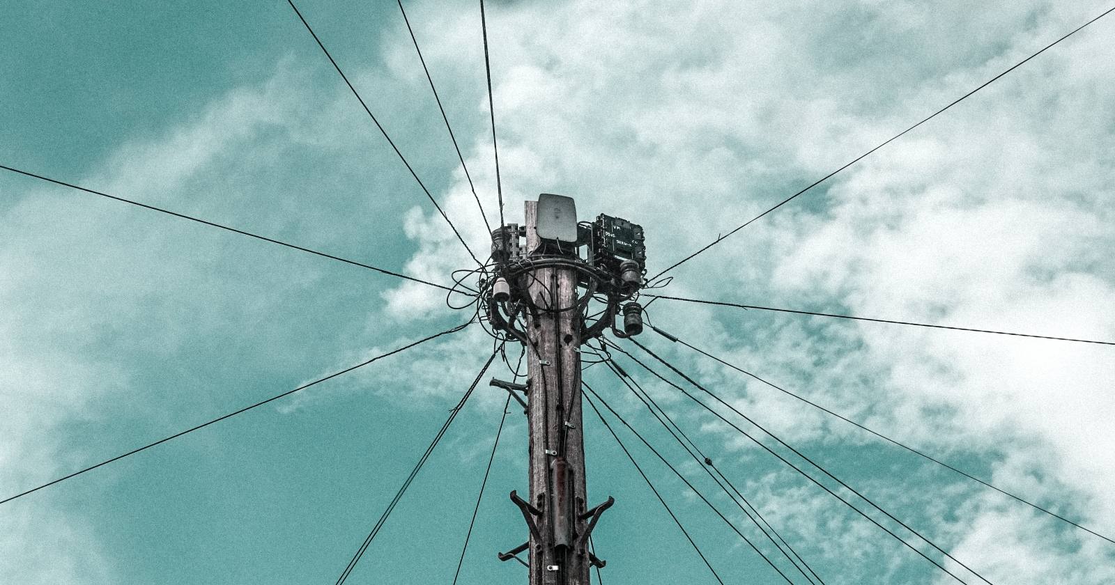 Poste de telecomunicaciones visto desde abajo, con cables que se abren hacia un cielo azul con nubes